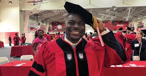 St. Johns Law alumnus Rashad Moore, wearing a red and black graduation gown and a black tam with a gold tassel, smiles with his hand lifted to his tam.